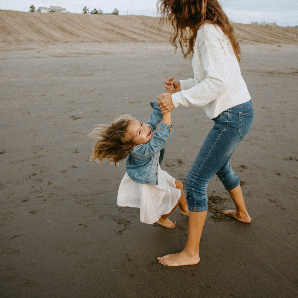 Une maman heureuse tient les mains de sa fille qui explose de rire sur la plage. On peut voir le sable doré en arrière-plan. La maman et sa fille portent toutes deux des tenues décontractées et assorties au bord de la mer.