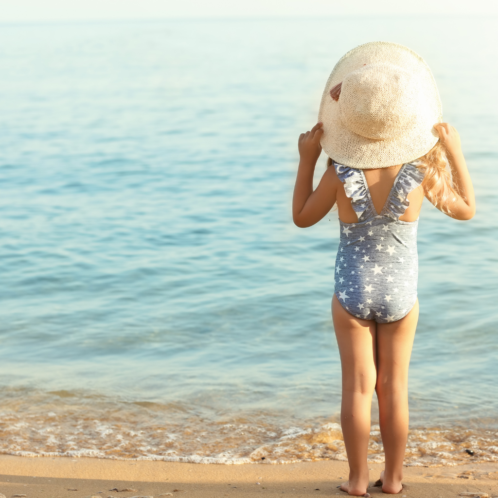 Une petite fille est debout de dos sur le sable, tenant un chapeau entre ses mains. Elle sourit, heureuse d'imiter sa maman. Le soleil brille et on peut voir l'océan à l'horizon. Le fond est rempli de couleurs chaudes et douces, créant une atmosphère chaleureuse et estivale.