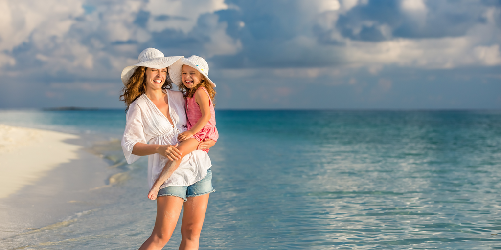 Image d'une maman qui porte sa fille sur le sable, toutes deux souriantes et heureuses. La maman tient sa fille dans ses bras et elles portent toutes les deux de jolis chapeaux. Le fond est composé d'un ciel bleu clair et d'une plage de sable doré.